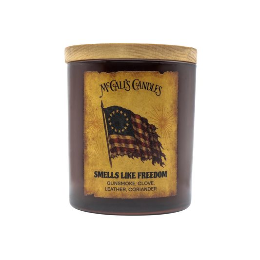 McCall's Candles Smells Like Freedom 8 oz amber jar candle with wood lid. Features distressed American flag and notes of gunsmoke, clove, leather, coriander.